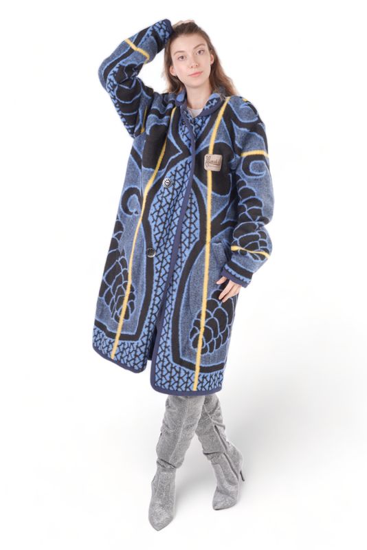 Female model in a blue patterned Basotho blanket jacket with yellow accents, hand on head, paired with shiny gray knee-high boots. She stands against a white background.