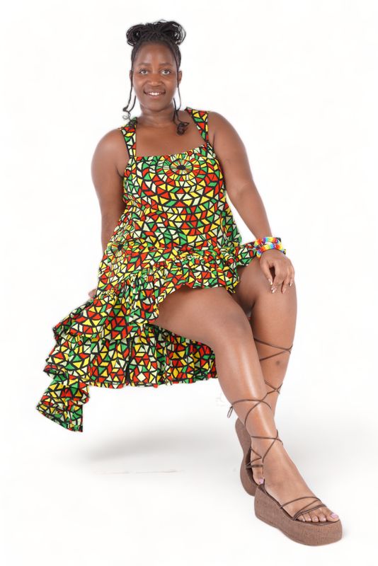 Woman wearing a vibrant, geometric-patterned high-low dress with red, yellow, and green colors. She accessorizes with a matching headband, earrings, and bracelets, showcasing a joyful, confident expression.