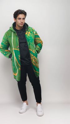 Man in a vibrant green coat with orange patterns, black pants, and white sneakers, stands confidently against a plain light background.