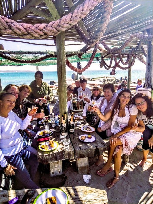 Eine Gruppe von Cap Classique enthusiasten prosten sich an einem langen rustikalen Holztisch im Freien direkt am Strand mit dem Meer im Hintergrund. ( Westkueste, Langebaan, Suedafrika.