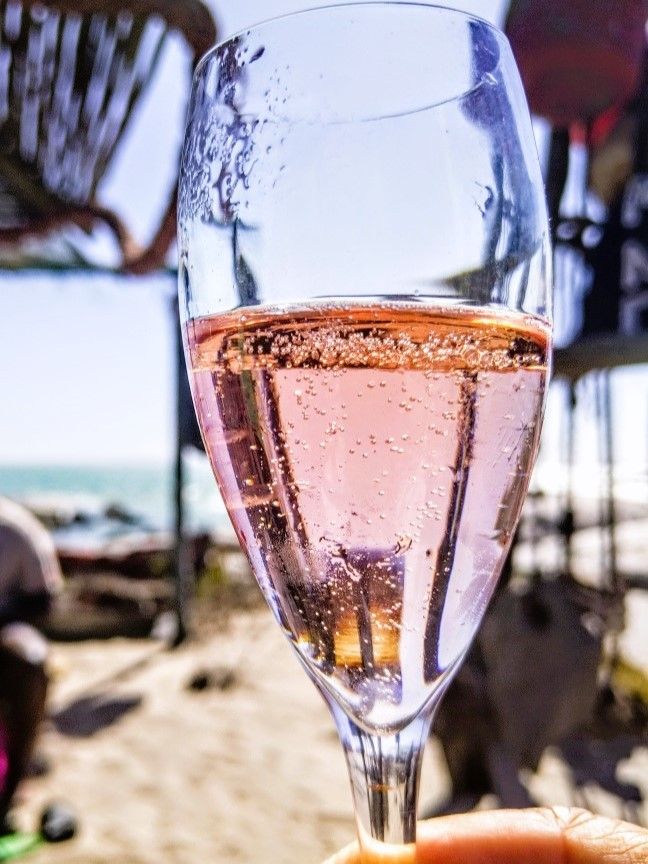 Ein Glas von Perlengefuellten Rose&#39; Cap Classique  wird in die Luft und gegen das Licht und den Ozean gehalten- lichtdurchflutet mit ganz kleinen, feinen Perlen. Langebaan, Suedafrika