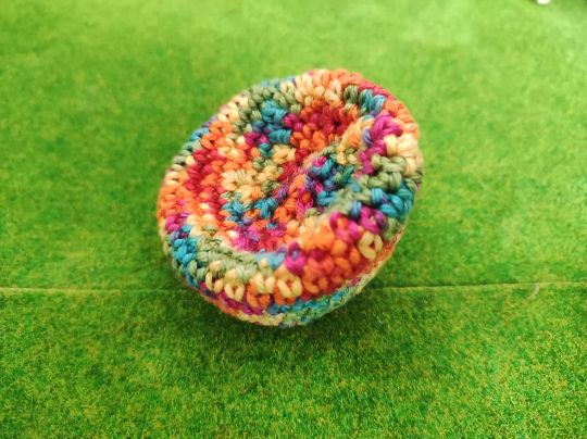 Handgehäkeltes Miniatur Sitzkissen mit buntem Farbverlauf auf einer grünen Deko Wiese. Farbverlauf in Rot, Orange, Gelb, Grün