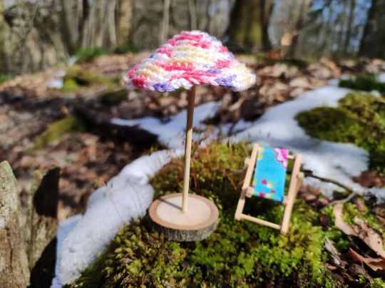 Gehäkelter Sonnenschirm mit Liegestuhl für die Wichtelwelt Gehäkelter Sonnenschirm mit Holzständer und Miniatur-Liegestuhl aus Holz mit blauem Bezug im Wald mit Moos und Schnee.