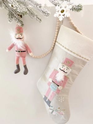 Mon Ami pink nutcracker Christmas ornament hanging from a evergreen branch with warm holiday lighting in the background