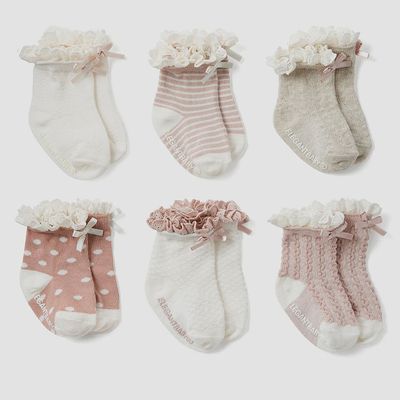 Six pairs of baby pink socks with ruffled cuffs and bow details arranged in a studio display, non-slip grip visible on the sole.