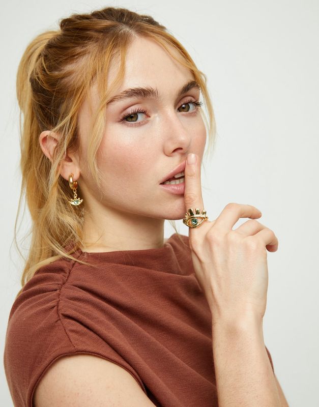 Gold eye-shaped ring with green crystal on a hand in natural light, showcasing the sculptural design and metalwork detail