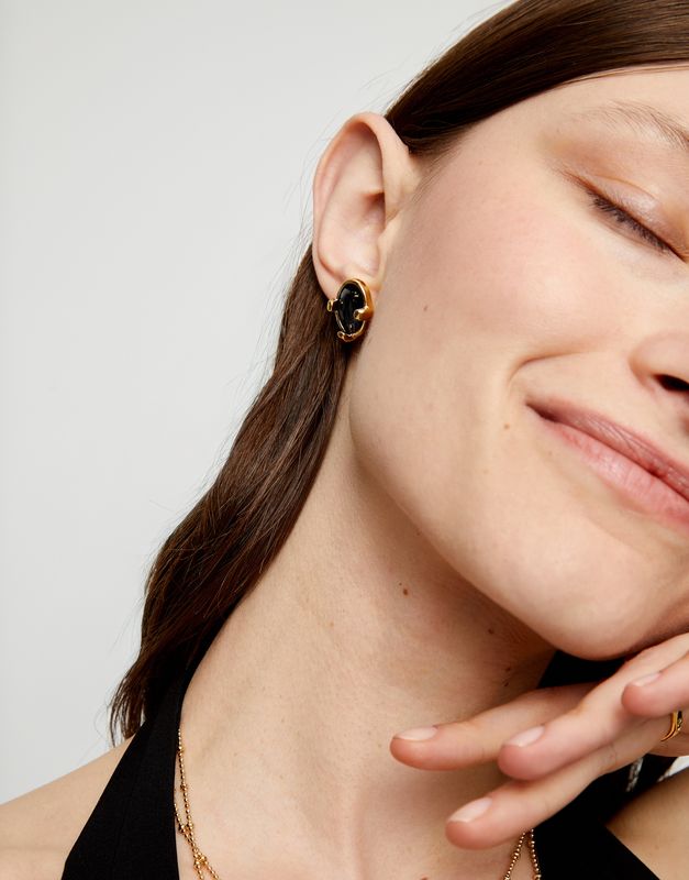 Jet black oval crystal stud earrings from Uno de 50's Ser Deslumbrante collection worn on a person, showing the polished stone and metal setting detail