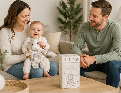 Baby sitting with mother and father holding giraffe plush lovey while gift box sits on table in cozy living room