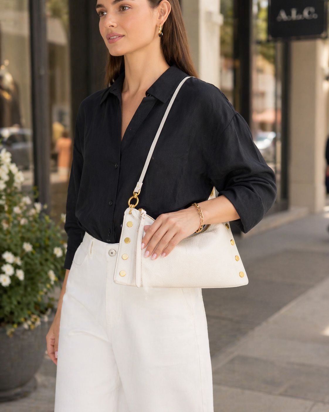 Woman in shopping district wearing black blouse and white pants with white Hammitt crossbody bag and gold hardware