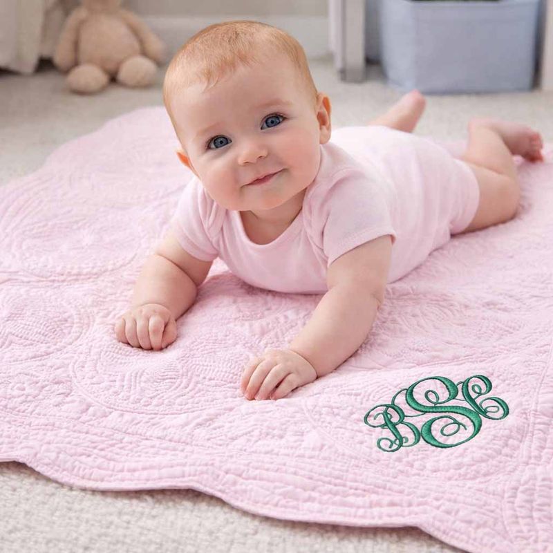 Baby lying on a personalized pink quilt with green embroidered monogram, soft quilted infant blanket in nursery setting