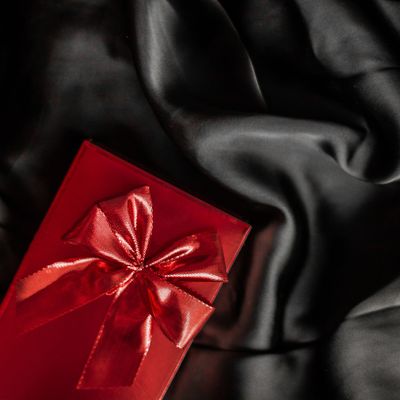 luxury gold and red gifting box from my geisha UK fragrance gifts for women. A vibrant red gift box adorned with a shimmering red ribbon, set against a backdrop of luxurious, dark satin fabric. The stark contrast between the bold red and the deep black evokes a sense of sophistication and mystery. The composition is simple yet elegant, drawing the viewer&#39;s eye to the meticulously tied bow, hinting at a special occasion or a carefully chosen gift. The use of rich fabrics and contrasting colors suggests luxury and indulgence, making it ideal for themes related to celebration, romance, or special occasions.