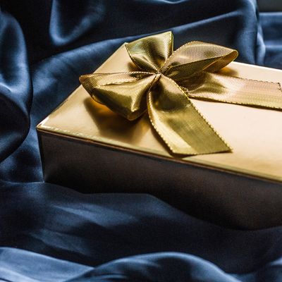 luxury golden gifting box on blue silk background from my geisha UK gifts for him. This image showcases a gold gift box adorned with a meticulously tied gold ribbon, set against a backdrop of luxurious blue silk fabric. The composition exudes elegance, sophistication, and a sense of anticipation associated with receiving a special gift. The choice of gold, often associated with wealth and prosperity, amplifies the feeling of luxury and importance attributed to the present. The contrasting dark blue silk background further accentuates the radiance of the gold, highlighting the gift as the central element and suggesting a sense of occasion. The image implies a moment of celebration or a gesture of deep affection, promising a treasured experience for the recipient. 