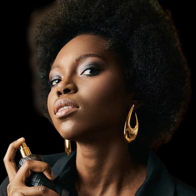 a woman with an afro hairstyle against a black background. The contrast emphasizes her features, illuminated by what appears to be a studio light. She holds a bottle of my Geisha perfume, about to spray, gaze fixed off-camera. This suggests a narrative, perhaps capturing her just before a night out. Her bold gold earrings and makeup suggest a confident, self-assured personality. The image speaks to themes of beauty, confidence, and allure. undefined
