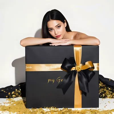 A woman rests her arms on a large black my Geisha gift box with a gold ribbon and bow, sitting on a white surface 