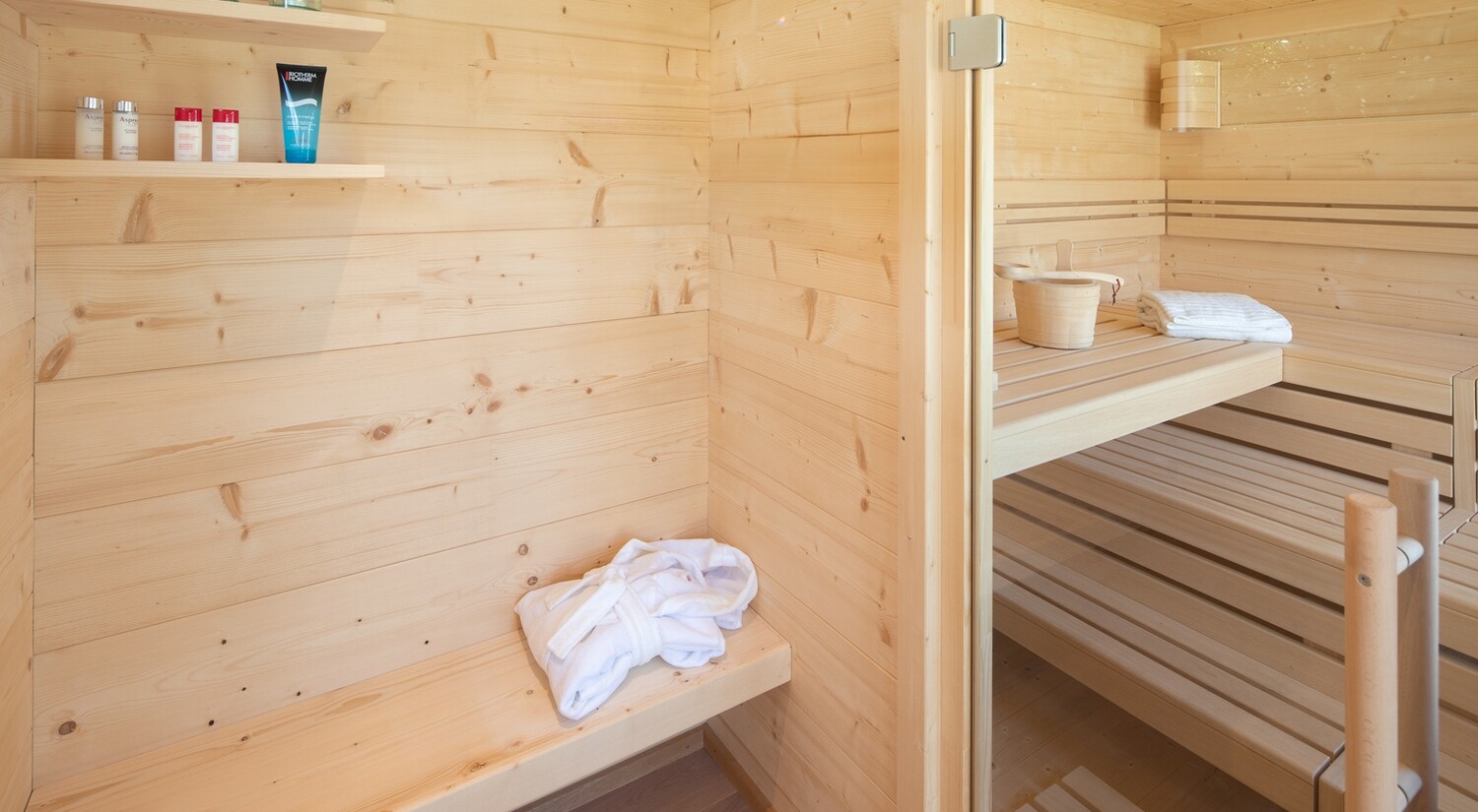 Innenraum mit großzügiger Glasfront, edler Fichtenholzverkleidung und ergonomischen Liegeflächen – schafft ein luxuriöses, lichtdurchflutetes Saunaerlebnis.