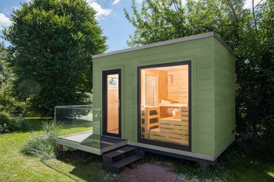 Modernes Saunahaus mit Flachdach und grüner Komodo-Lasur, klare Kubusform und Holz-Aluminium-Fenstern – ein stilvoller Rückzugsort für den Garten.​