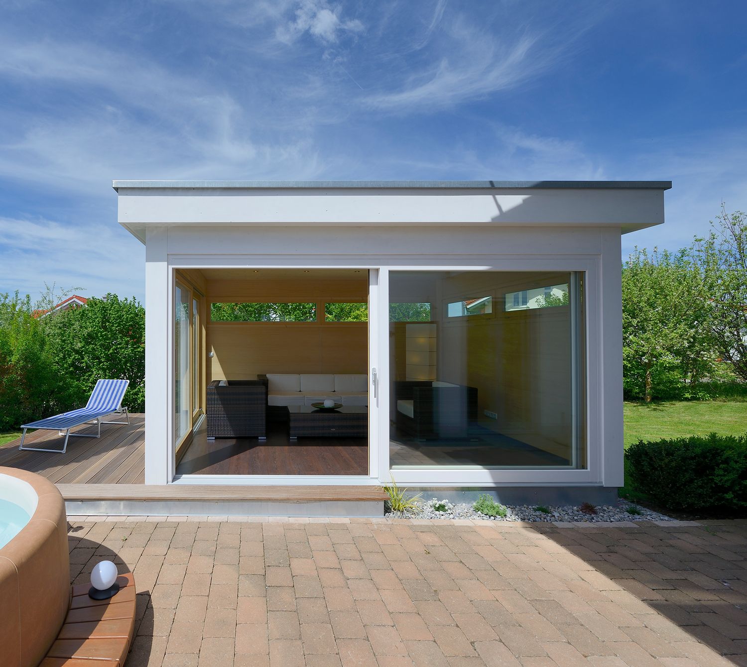 Modernes Gartenhaus mit weißem Flachdach und großzügigen Hebe-Schiebetüren – minimalistisches Design im Bauhaus-Stil, harmonisch eingebettet in eine grüne Gartenlandschaft.