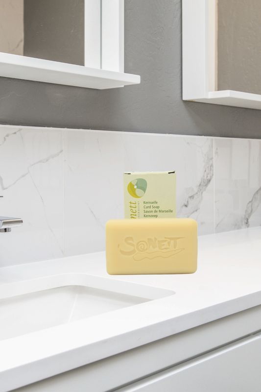 Sonett Kernseife 100 g auf weißer Marmoroberfläche mit Zahnbürste & Glasbehälter im Hintergrund, beige Seifenstück mit Sonett