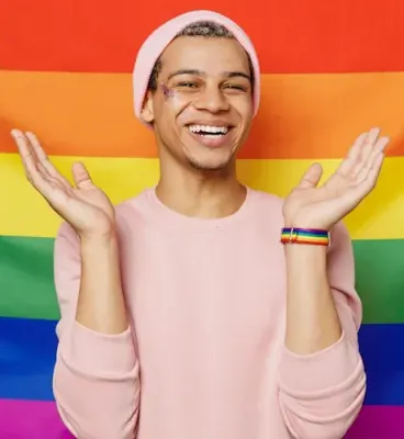 Zeig deine Pride mit unserer neuen Kollektion! Junger Mann mit Pride-Armband vor Regenbogenflagge. Entdecke jetzt unsere Armbänder.