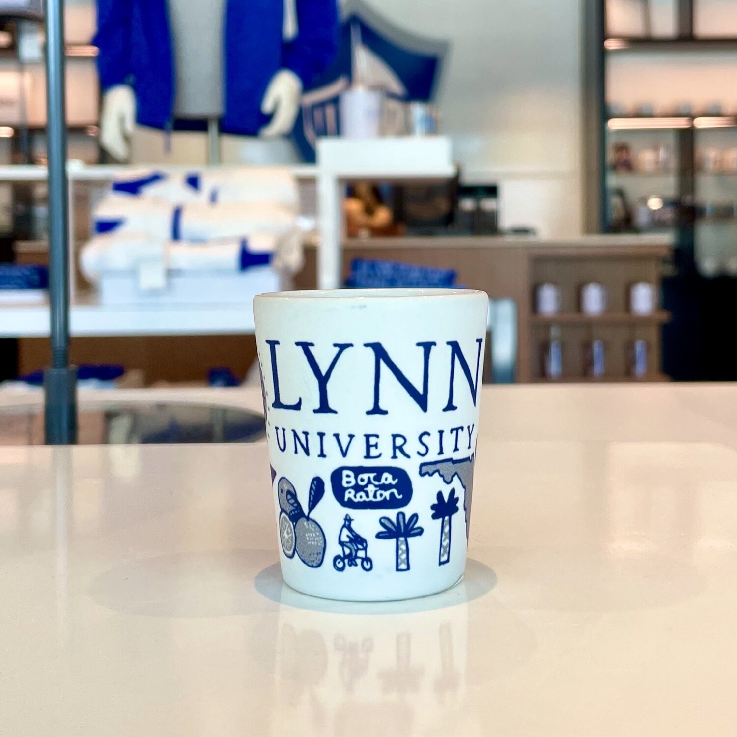 White ceramic shot glass  with blue Lynn logos and blue text saying &quot;Lynn University.&quot;