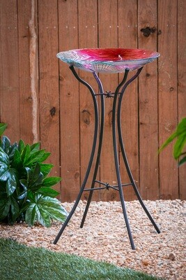 Metal bird bath stand holding a colorful glass bowl in a garden setting, surrounded by green foliage.