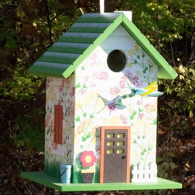 The Grasshopper Birdhouse hanging from a tree branch in a sunlit garden, surrounded by green leaves and white flowers.