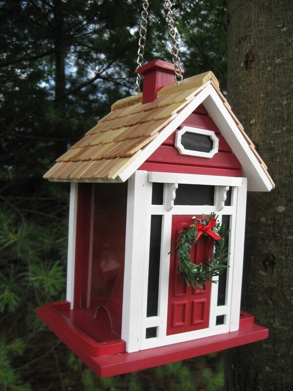 White cottage bird feeder with red trim, a pine shingled roof, and a green holiday wreath above a red door.