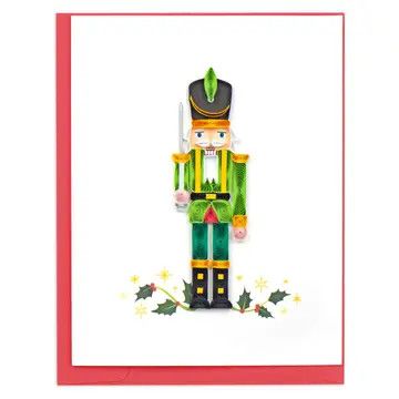 Quilled paper nutcracker in a red jacket and green pants on a white card with holly and gold stars.