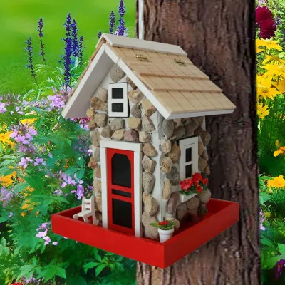 Stone guest cottage bird feeder hanging from a tree branch in a garden setting, showcasing the pine-shingled roof.