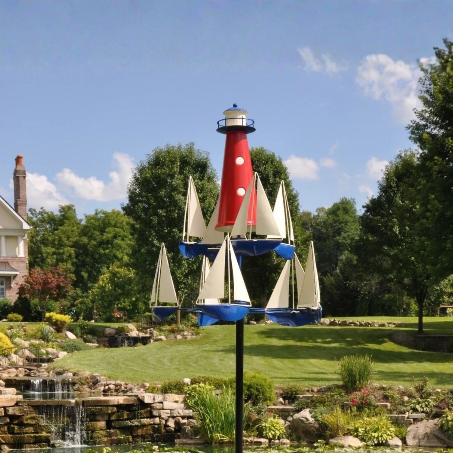 Metal lighthouse and sailboat nautical wind spinner on tall black garden stake displayed in landscaped yard.