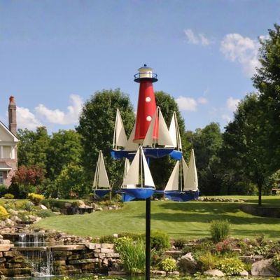 Metal lighthouse and sailboat nautical wind spinner on tall black garden stake displayed in landscaped yard.