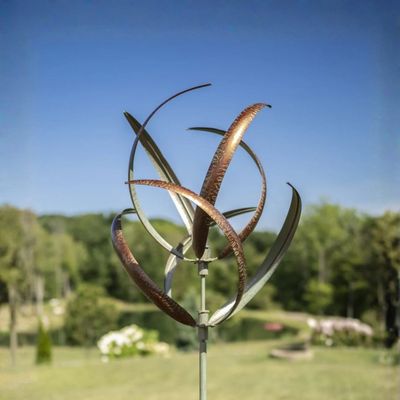 Copper and verde metal wind spinner with curved dual spinning blades mounted on garden stake outdoors.