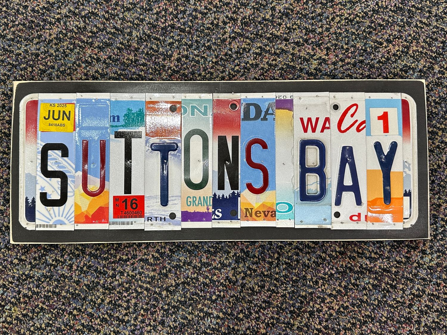 Rectangular wooden sign displaying the words "SUTTONS BAY" spelled out using cut pieces of colorful upcycled vintage licenses