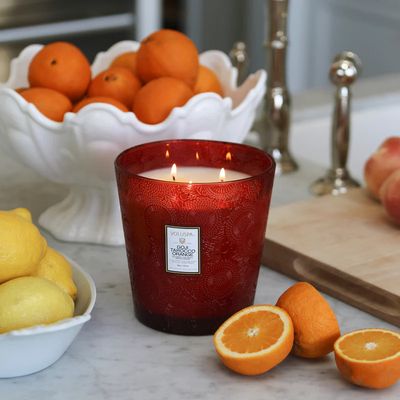 Large three-wick candle in a vibrant red embossed glass jar alongside a bowl of fresh oranges