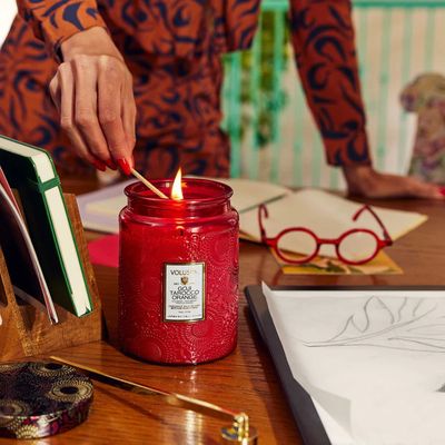 Lifestyle shot of a lit Goji Tarocco Orange candle next to a glass of citrus-infused water, showcasing the warm ruby glow of 