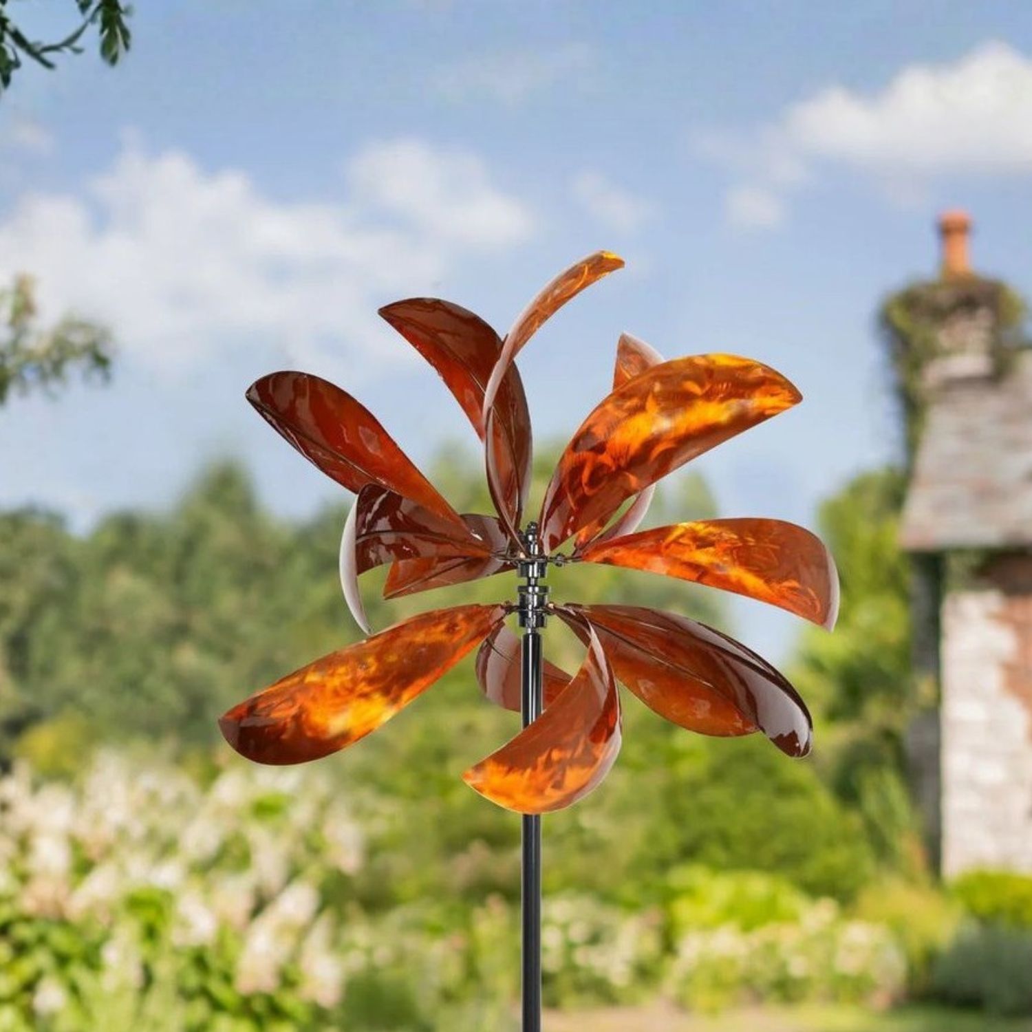 Bronze metallic wind spinner with curved sculptural blades mounted on tall black garden stake in landscaped yard.