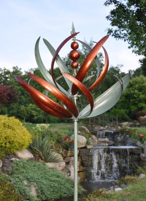 Copper and verde metal wind spinner with curved reed-like blades mounted on tall garden stake in landscaped yard.