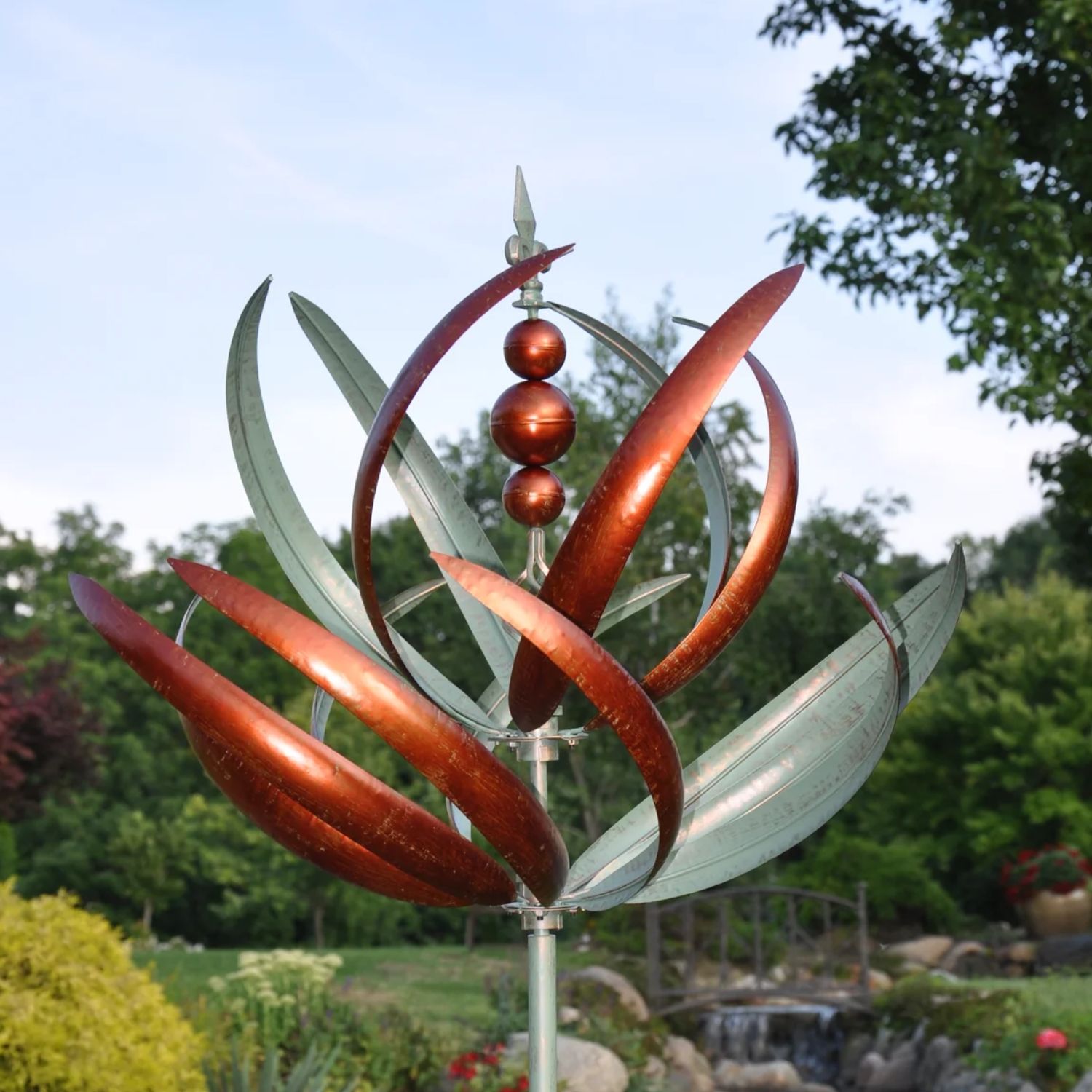 Copper and verde metal wind spinner with curved reed-like blades mounted on tall garden stake in landscaped yard.
