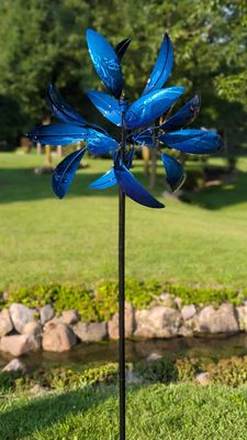 Blue Luminous Windemere metal wind spinner with layered curved blades on tall garden stake in landscaped yard.