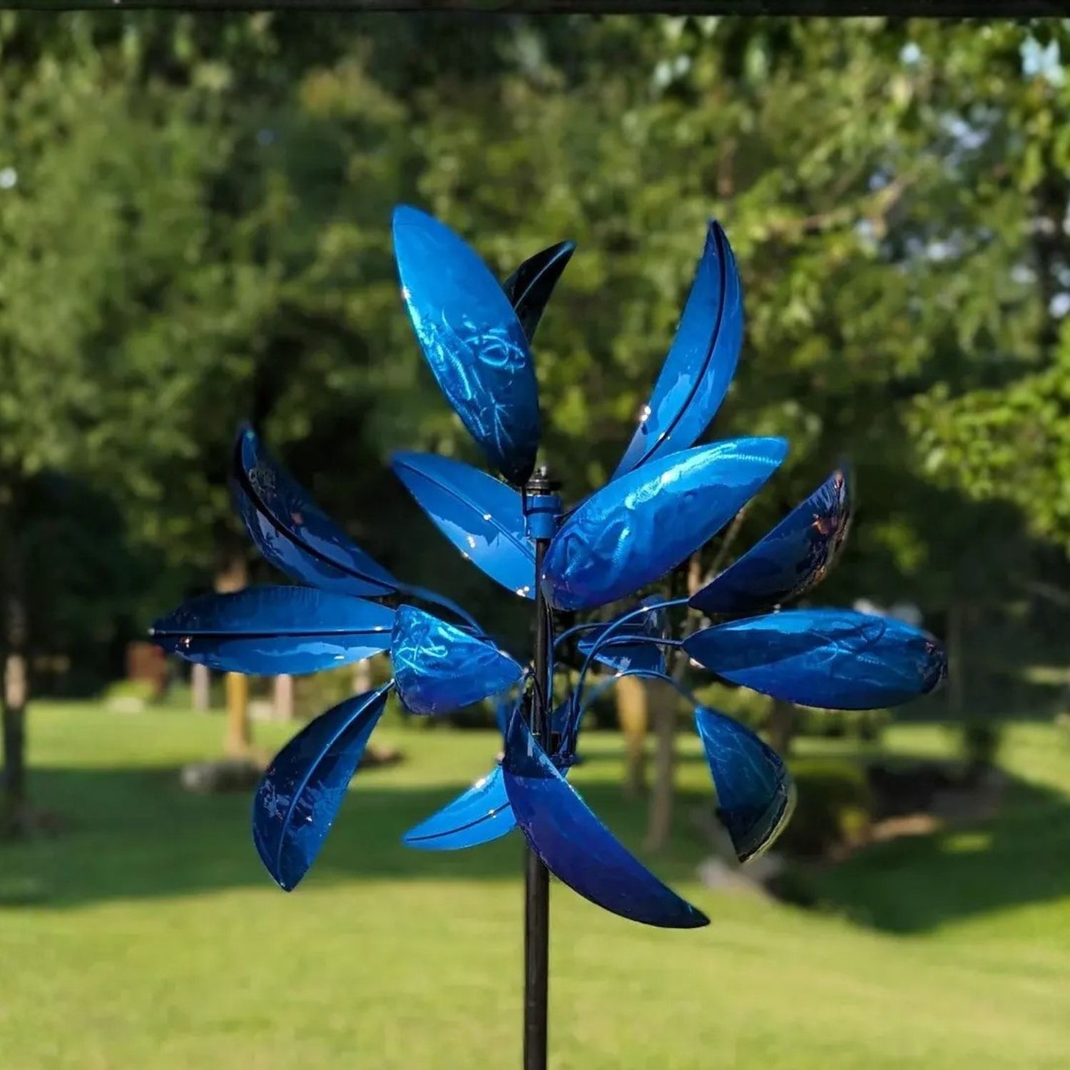 Blue Luminous Windemere metal wind spinner with layered curved blades on tall garden stake in landscaped yard.