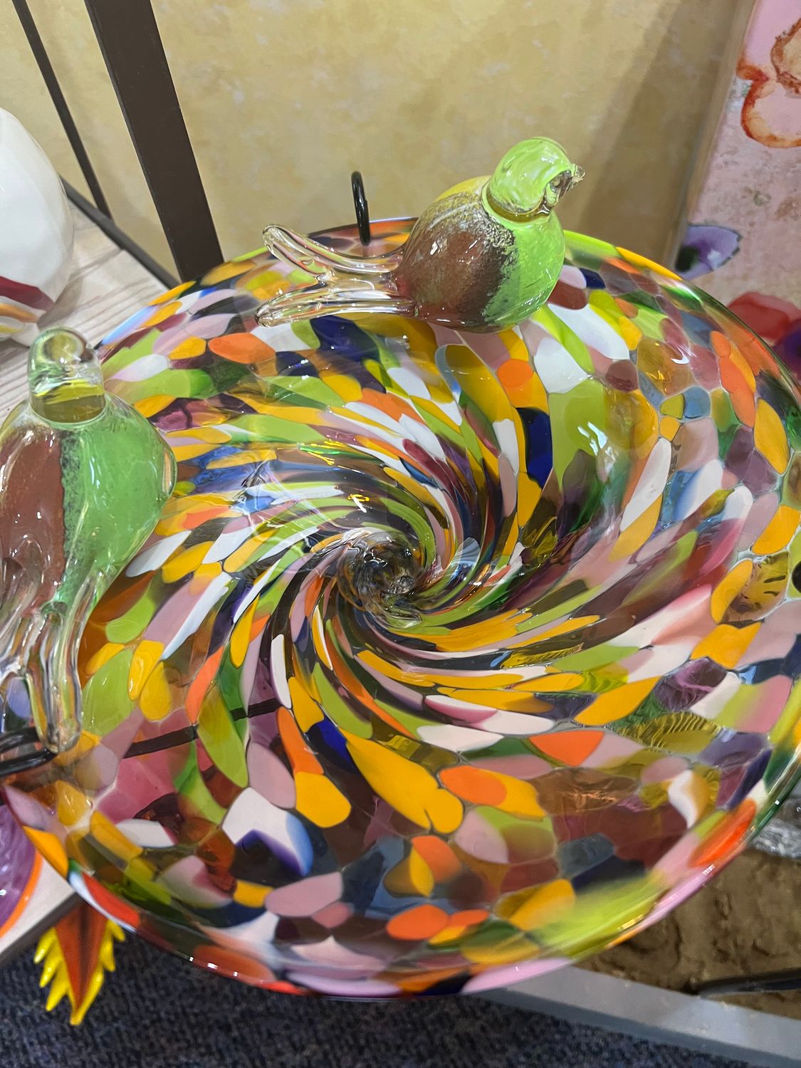 Close-up top view of a colorful multi-opaque glass birdbath bowl showing an intricate swirling pattern of yellow, red, blue