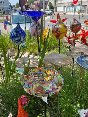 Glass birdbath with vibrant multi-colored opaque pattern mounted on a black metal stand in a garden setting next to plants