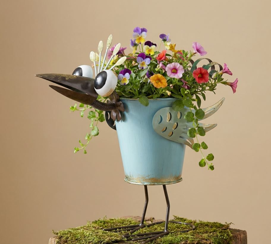 Colorful turquoise metal peacock planter with oversized eyes, decorative wings and curled tail feathers filled with flowers.