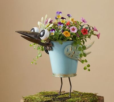 Colorful turquoise metal peacock planter with oversized eyes, decorative wings and curled tail feathers filled with flowers.