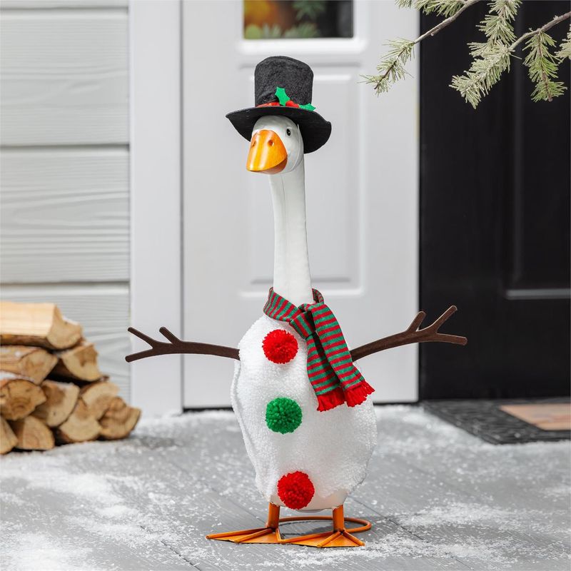 Giddy the Porch Goose wearing a white snowman outfit, striped scarf, and black top hat beside a front door and stacked wood.