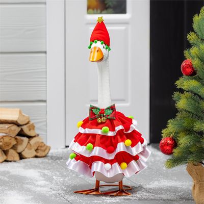 Giddy the Porch Goose wearing a red ruffled Christmas dress with colorful pom-poms and a star-topped hat beside an evergreen.