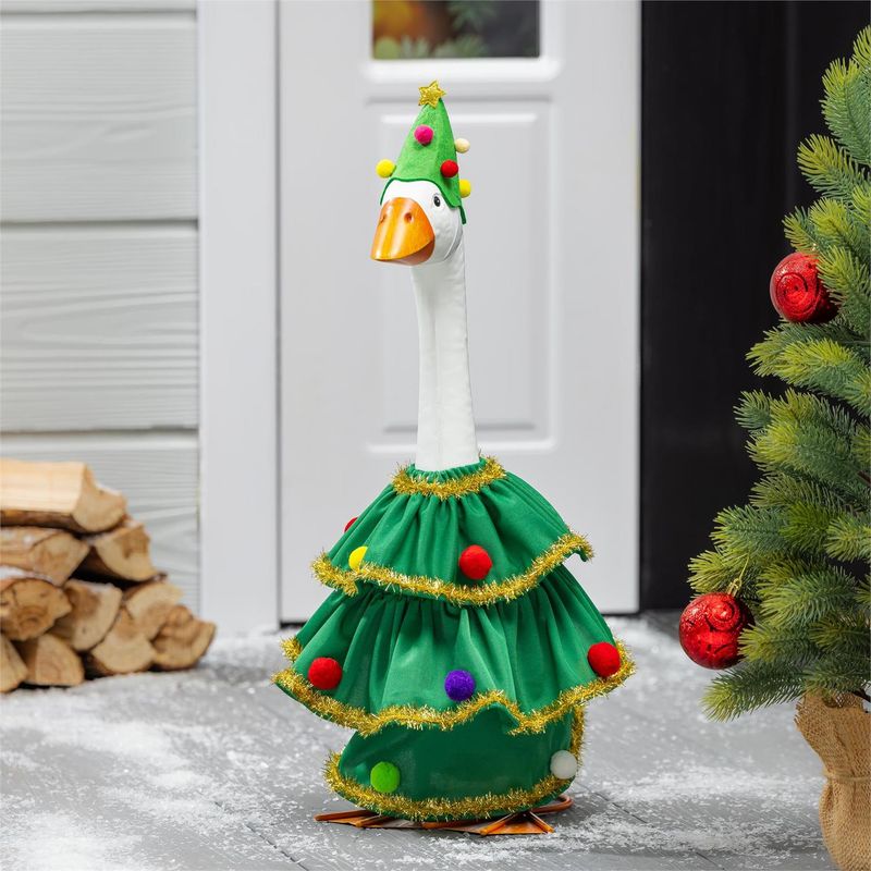 Giddy the Porch Goose wearing a green Christmas tree outfit with gold tinsel and colorful pom-poms beside a decorated evergre