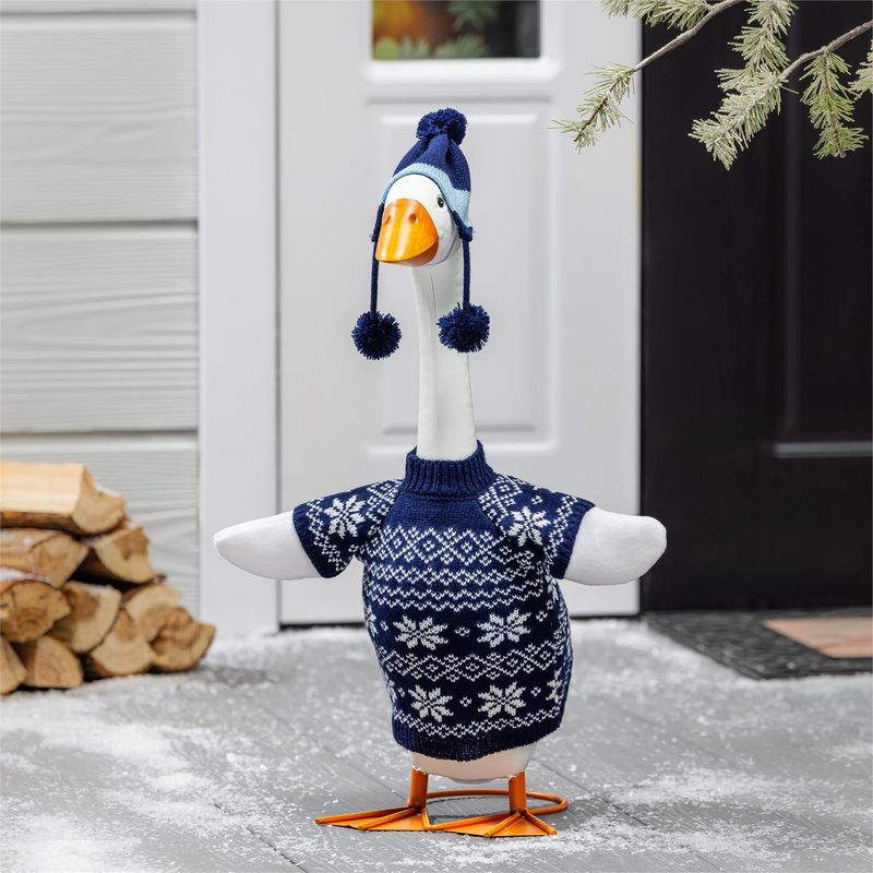 Giddy the Porch Goose in a navy snowflake sweater and pom-pom hat beside a front door and stacked firewood.