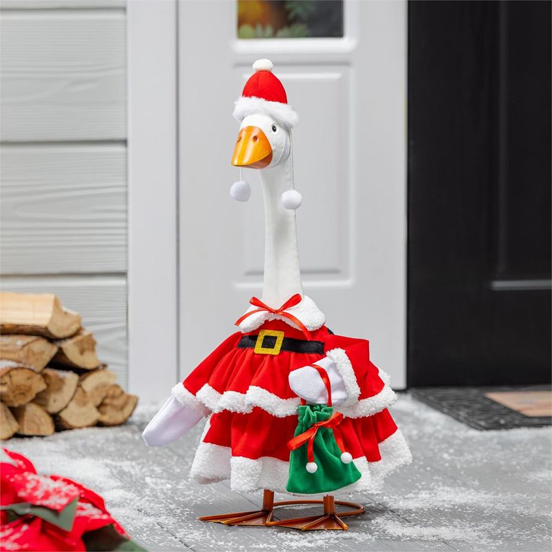 Giddy the Porch Goose wearing a red Mrs. Claus dress and pom-pom hat with a green gift bag beside a winter entryway.