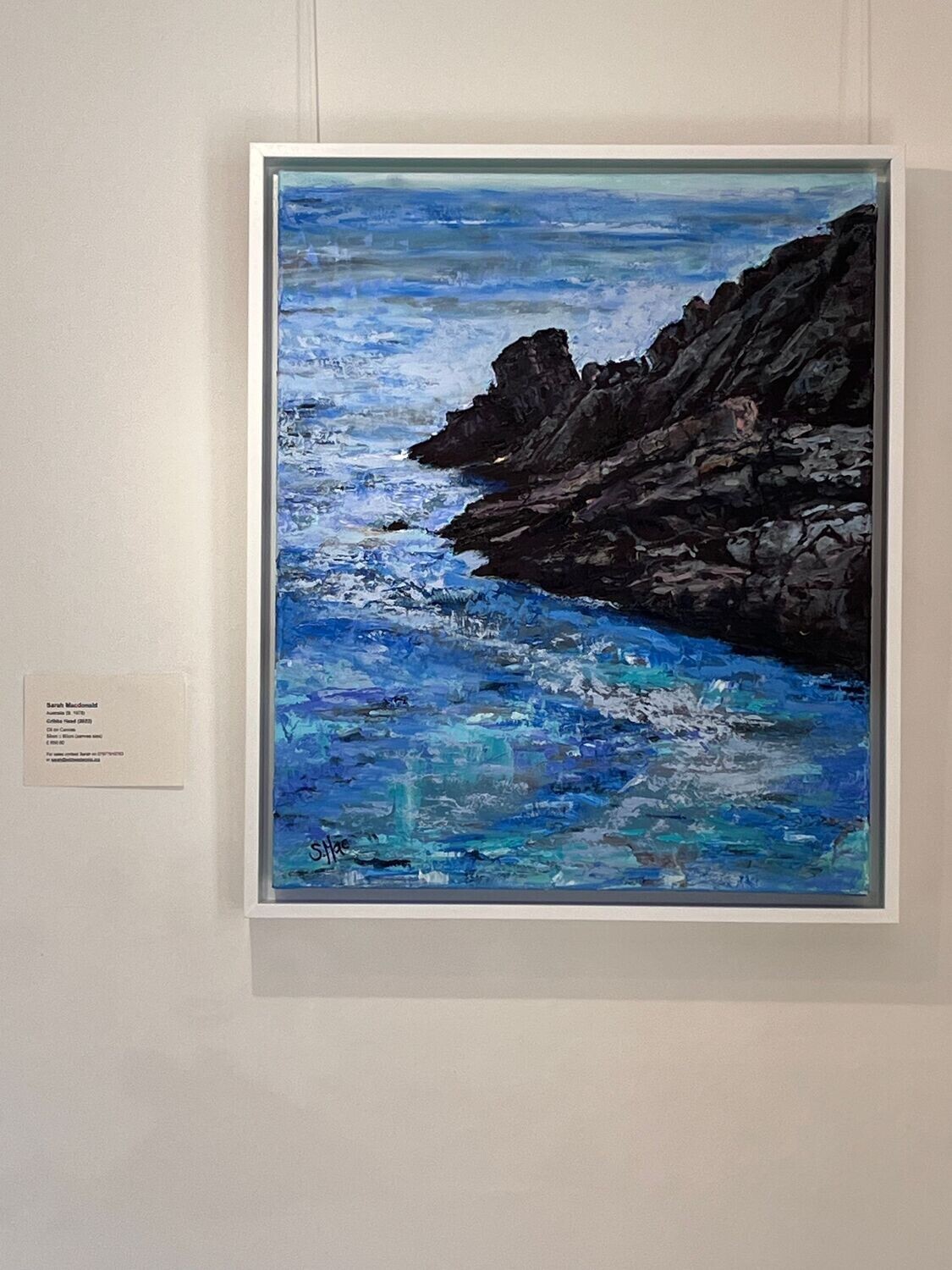 framed portrait orientated painting with label to the left. The painting is two thirds sea with one third being rocky headland. The rocks are dark brown, peach and grey, the sea is vibrantly textured aqua, blues, greys and lavender. The sea wraps around the rocks which are coming out of the frame from the top right.